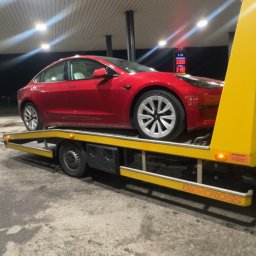 JOKER - Czerwona Tesla Model 3 na żółtej lawecie pod zadaszeniem stacji benzynowej, widok z boku, nocne oświetlenie.