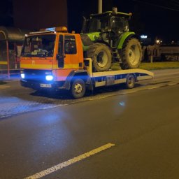 JOKER - Pomarańczowa laweta z niebieskimi światłami przewozi zielony traktor w nocy. Widoczny numer rejestracyjny lawety i oświetlenie uliczne w tle.