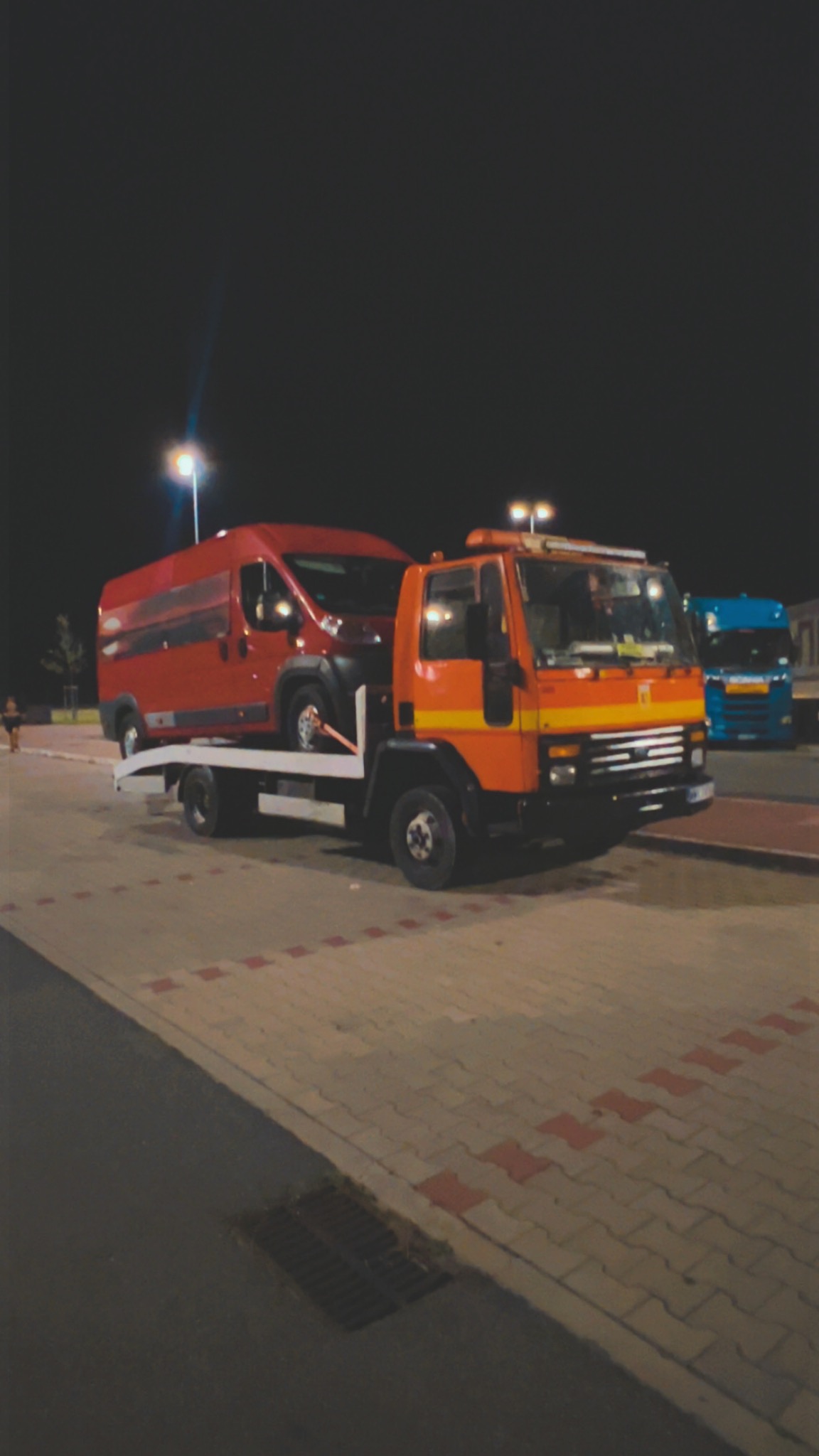 Pomarańczowa laweta z czerwonym busem na platformie, nocą, na parkingu z kostki brukowej. W tle widoczny niebieski samochód ciężarowy.