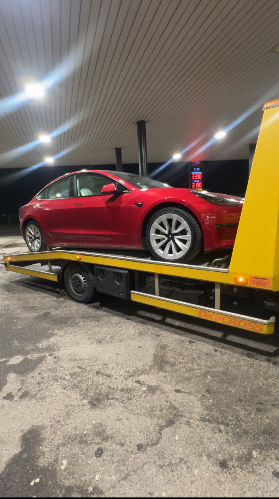 Czerwona Tesla Model 3 na żółtej lawecie pod zadaszeniem stacji benzynowej, widok z boku, nocne oświetlenie.