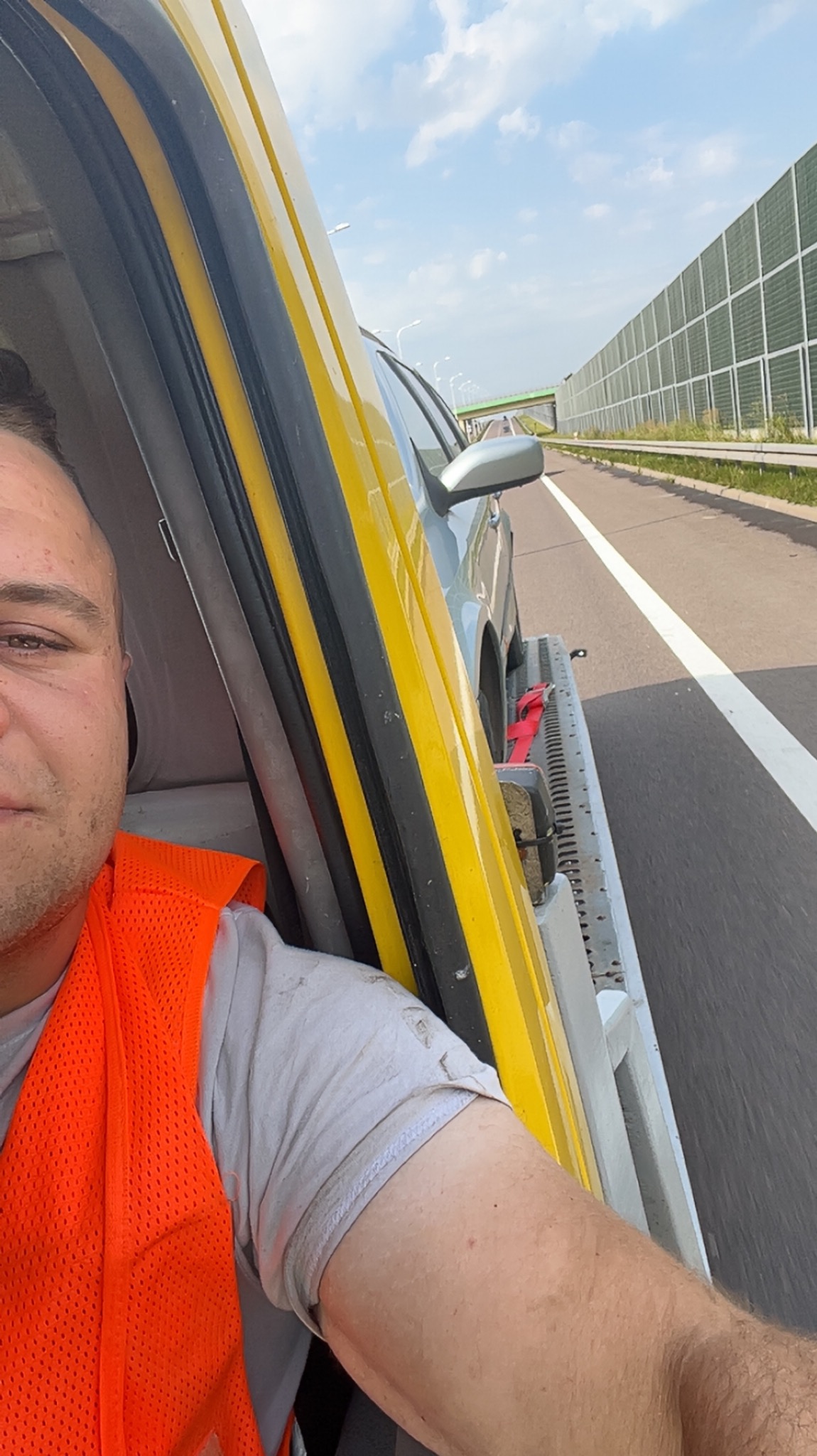 Laweta w ruchu: widok z wnętrza kabiny, pomarańczowa kamizelka pracownika, przewożony samochód na platformie, autostrada z barierami akustycznymi.