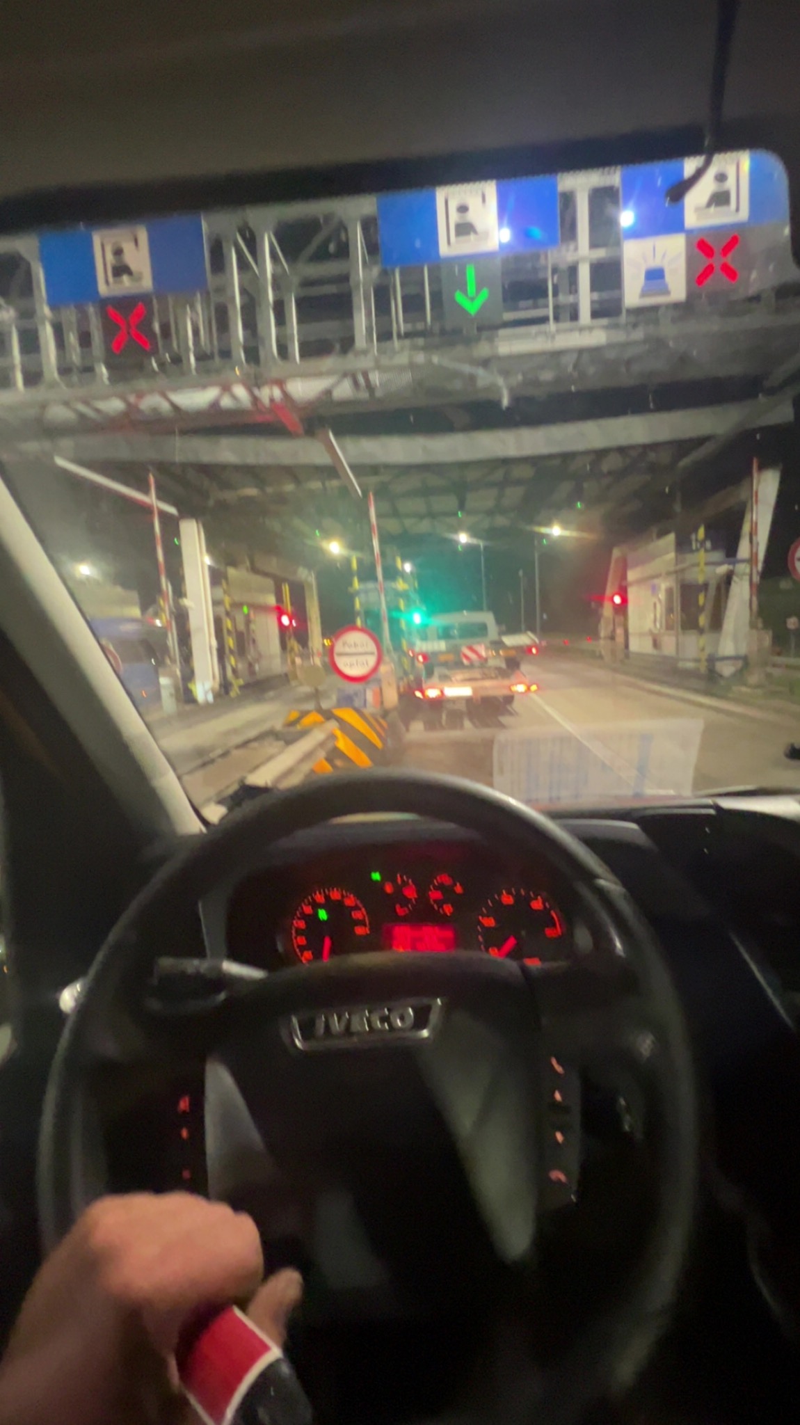 Widok z kabiny Iveco na punkt poboru opłat drogowych nocą. Nadjeżdżający samochód ciężarowy z ładunkiem. Czerwone i zielone światła nad bramkami.