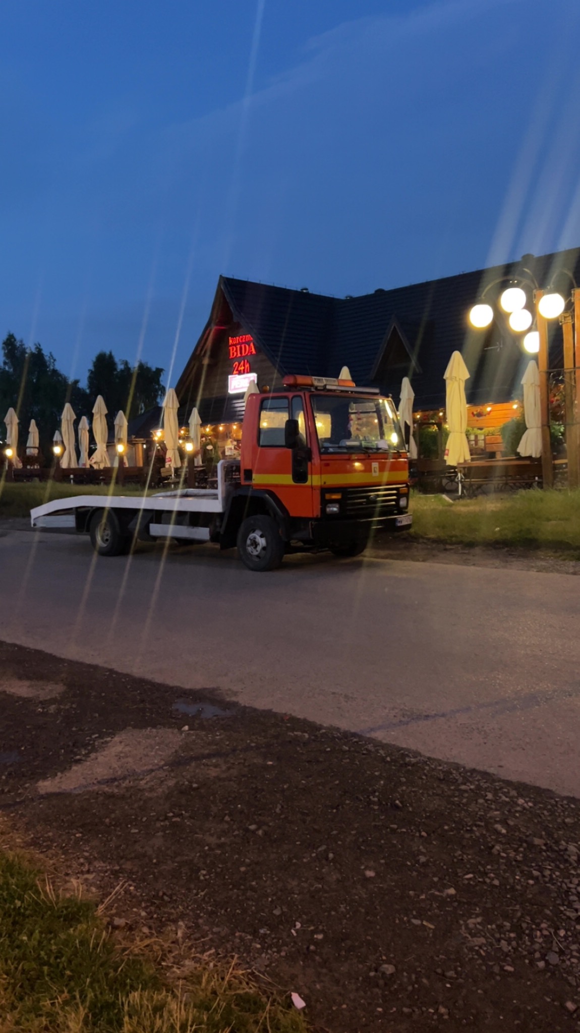 Pomarańczowa laweta stoi na drodze przed oświetlonym budynkiem z neonem 'BIDA 24h' na dachu, wieczorne niebo w tle.