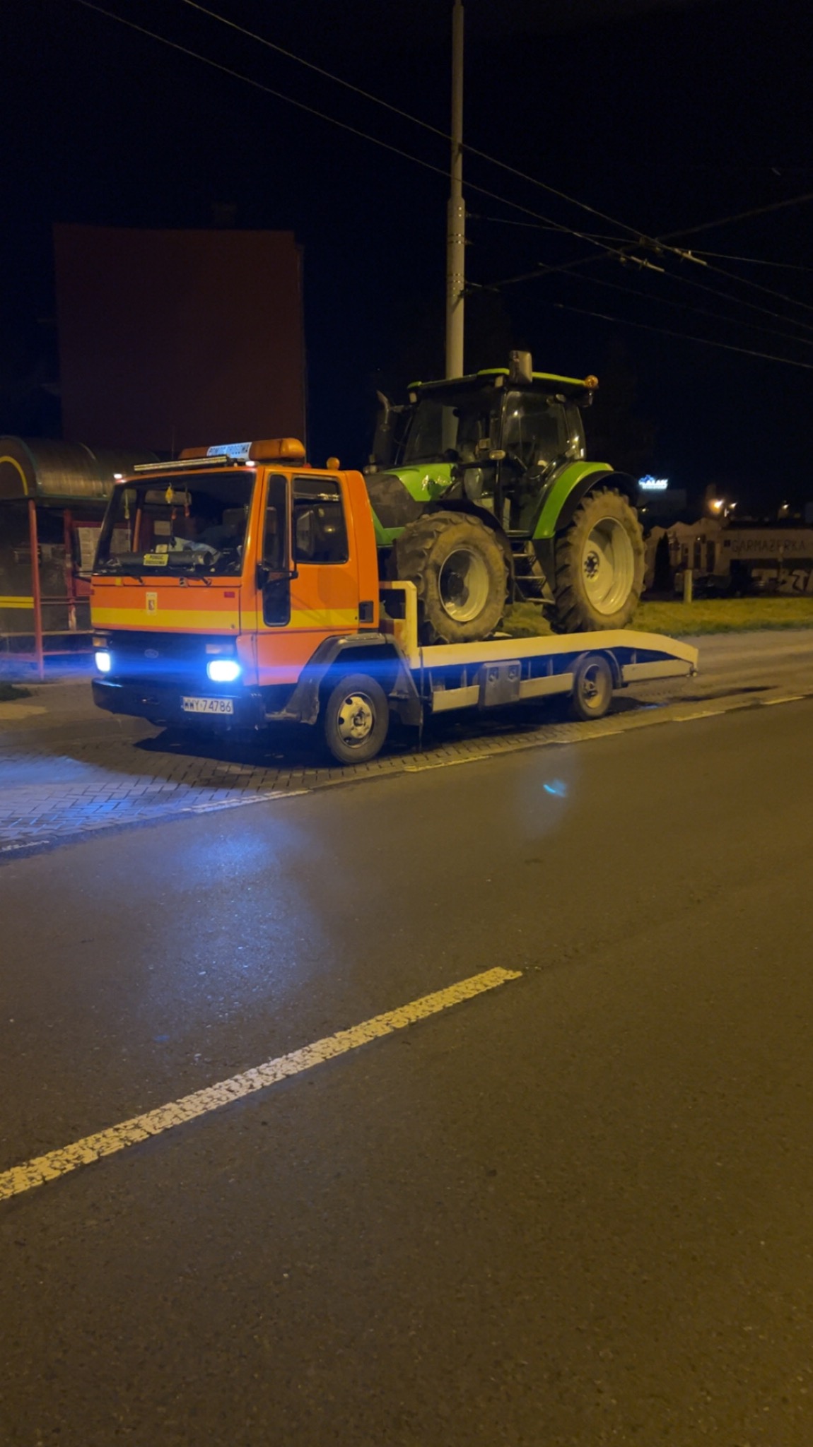 Pomarańczowa laweta z niebieskimi światłami przewozi zielony traktor w nocy. Widoczny numer rejestracyjny lawety i oświetlenie uliczne w tle.