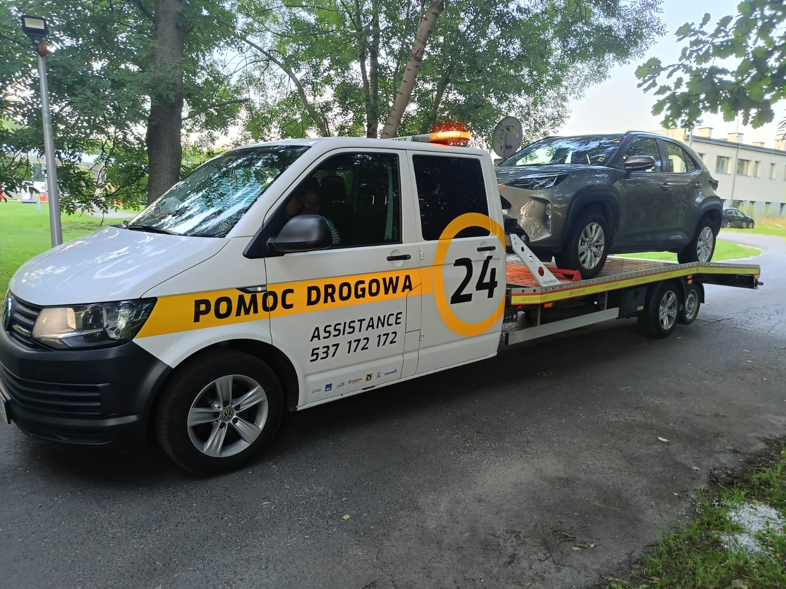 Laweta z szarą Toyotą Yaris Cross na platformie, zaparkowana na asfaltowej drodze w otoczeniu zieleni. Widoczny numer telefonu i napis 'Pomoc Drogowa'.