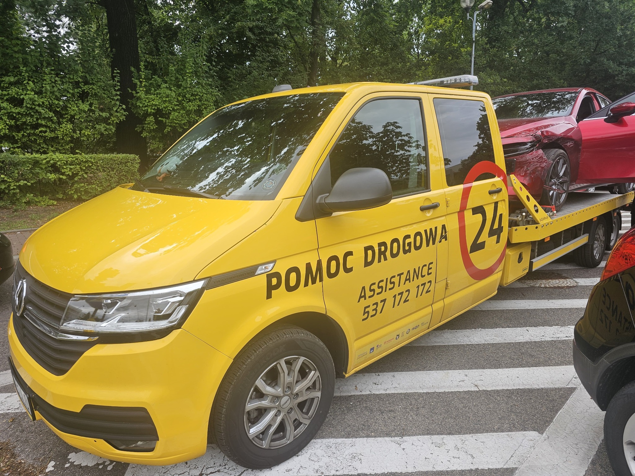 Żółta laweta Pomoc Drogowa z czerwonym uszkodzonym autem na platformie, zaparkowana na przejściu dla pieszych, zieleń w tle.