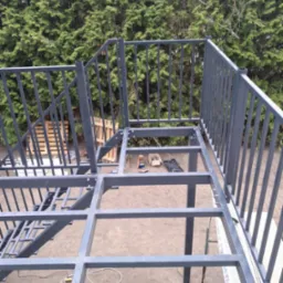 Metalowa konstrukcja schodów zewnętrznych z balustradą w trakcie montażu, z widocznym zarysem schodów i zielonym tłem z drzew.