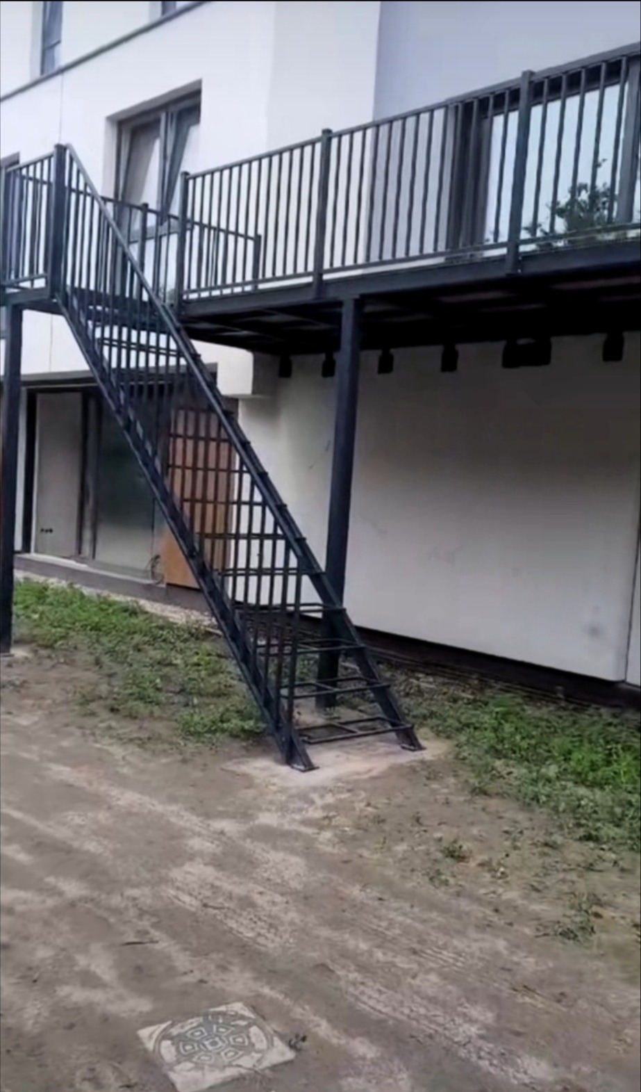 Zewnętrzne schody metalowe z balustradą prowadzące na balkon. Konstrukcja pomalowana na czarno, na zewnątrz budynku z białymi ścianami. Widok z dołu.