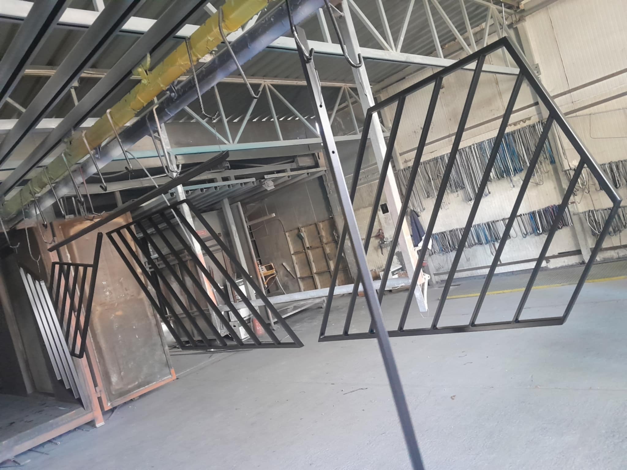 Malowane proszkowo, metalowe elementy ogrodzenia o ukośnym wzorze, wiszące na wieszakach w hali produkcyjnej. Widoczne elementy konstrukcji dachu.