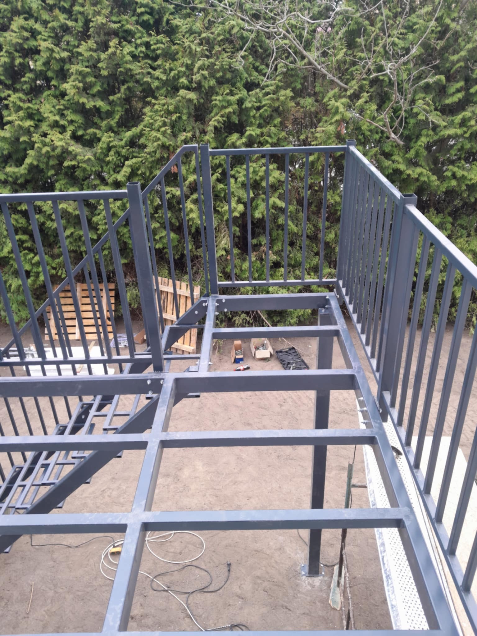 Metalowa konstrukcja schodów zewnętrznych z balustradą w trakcie montażu, z widocznym zarysem schodów i zielonym tłem z drzew.