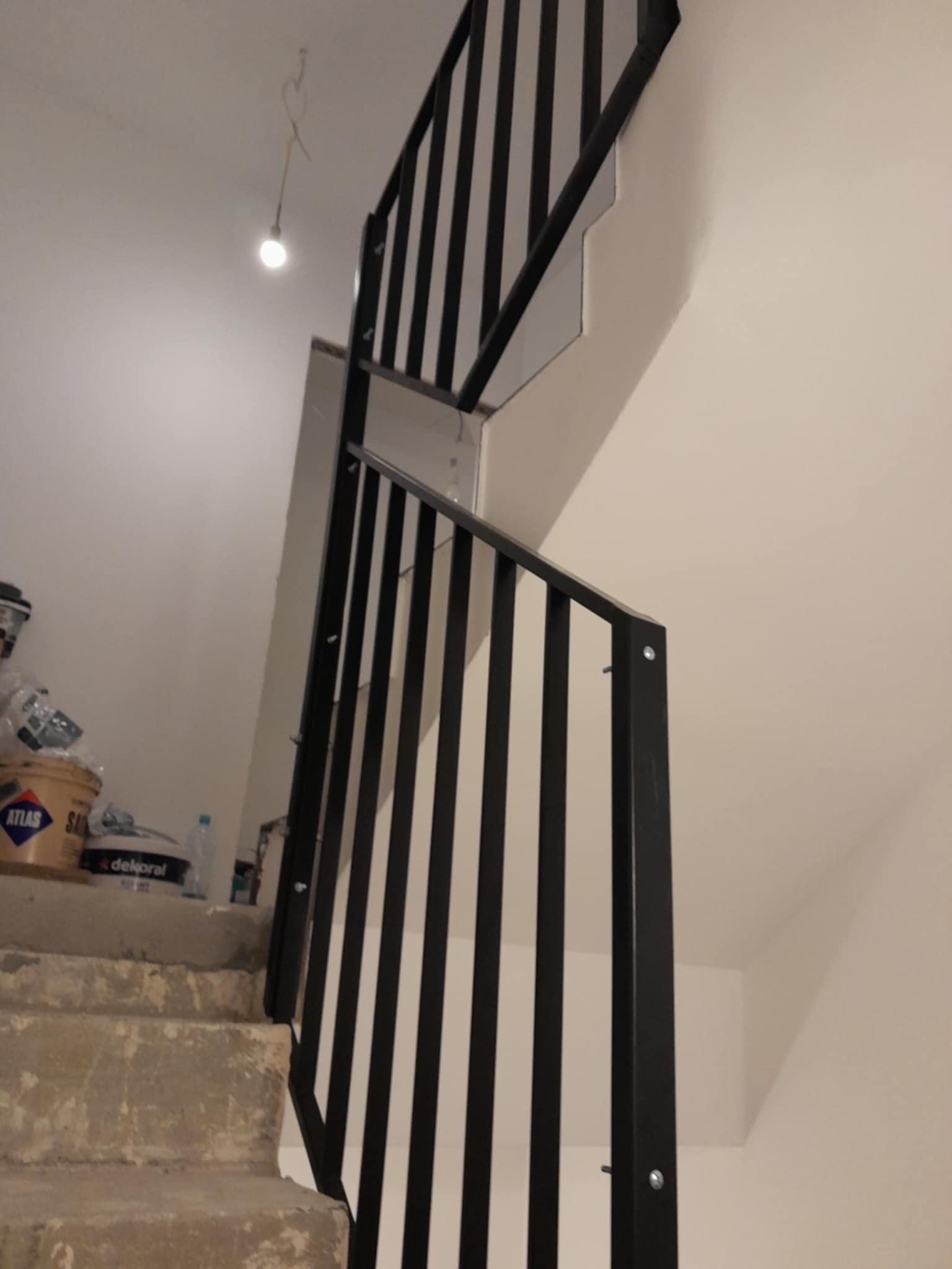 Nowoczesna, czarna balustrada metalowa z pionowymi szczeblami, zamontowana na betonowych schodach w jasnym wnętrzu, z widocznymi puszkami farb w tle.