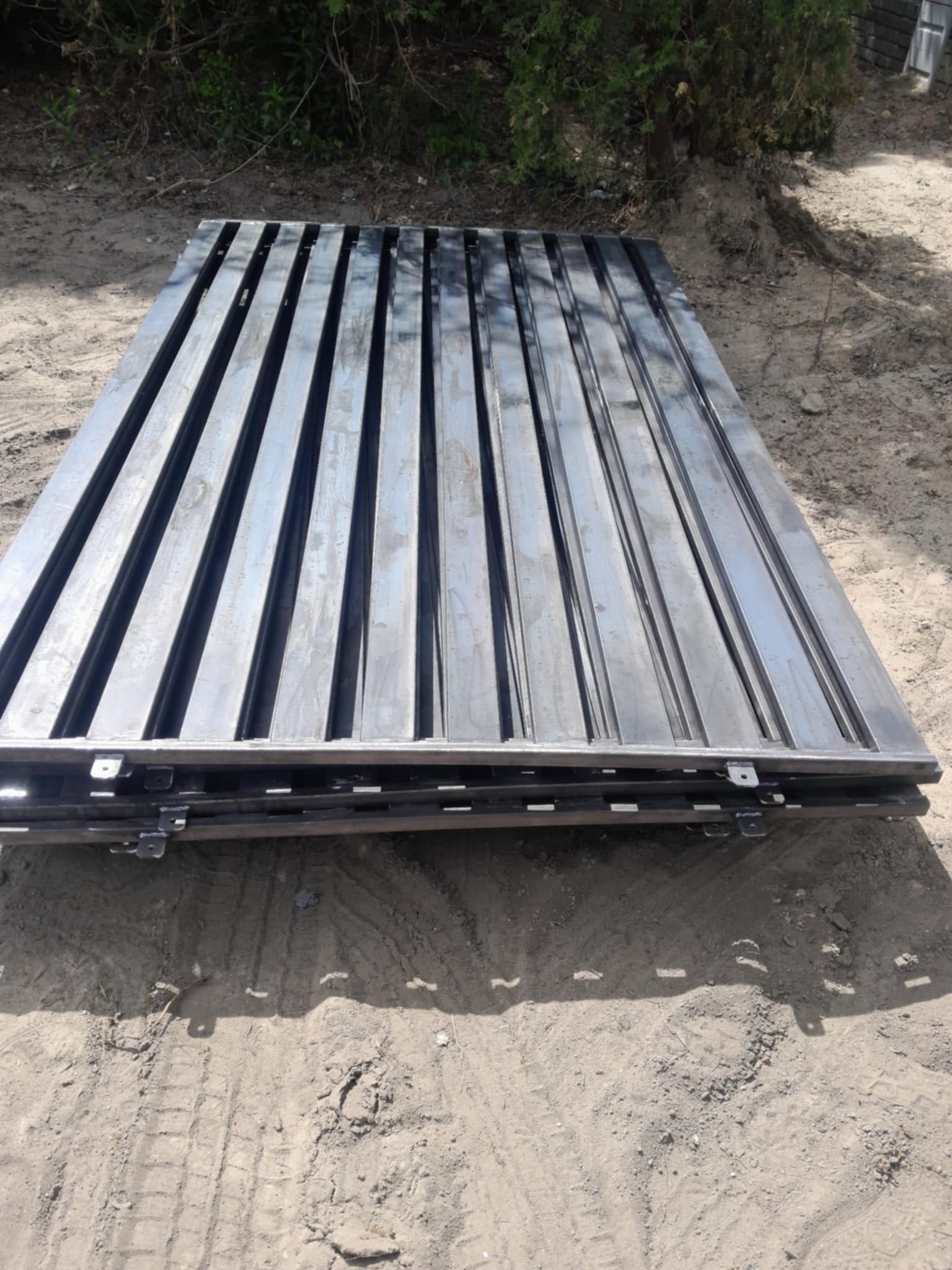 Stos paneli ogrodzeniowych z metalu, z widocznymi uchwytami montażowymi, leżący na ziemi. Panele w ciemnym kolorze, ułożone równo.