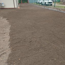 Tomek - Przygotowany teren pod trawnik z widocznymi śladami po maszynach, ogrodzeniem i budynkiem w tle. Grunt wyrównany, gotowy do siewu lub rozłożenia trawy z rolki.