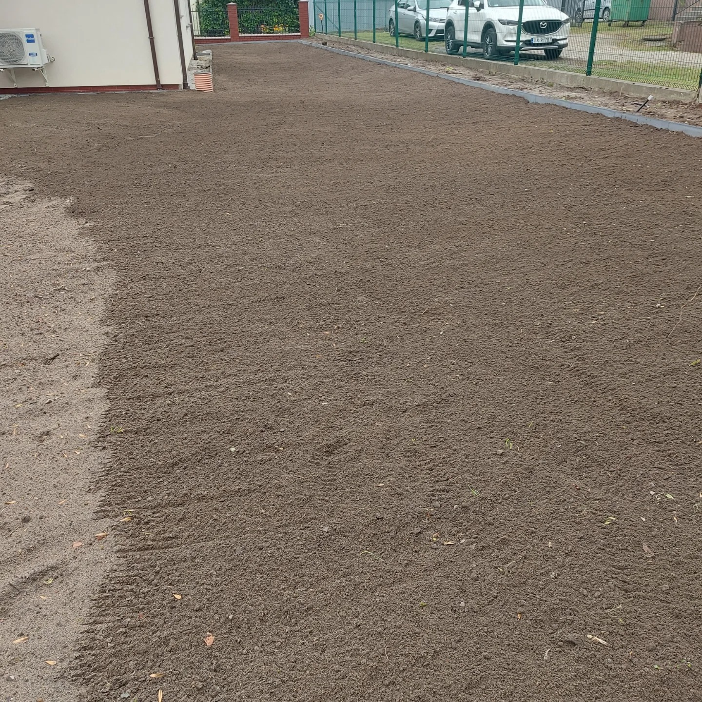 Przygotowany teren pod trawnik z widocznymi śladami po maszynach, ogrodzeniem i budynkiem w tle. Grunt wyrównany, gotowy do siewu lub rozłożenia trawy z rolki.