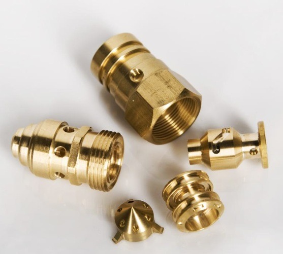 Precyzyjne, mosiężne elementy z gwintami, różnej wielkości i kształtu, leżące na białym tle. Widoczne detale obróbki, otwory i gwinty.