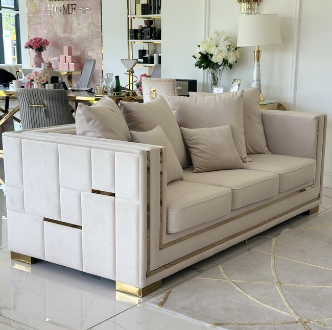Beżowa sofa z geometrycznym wzorem na bokach i złotymi akcentami, ustawiona w eleganckim salonie z dekoracjami w stylu glamour.