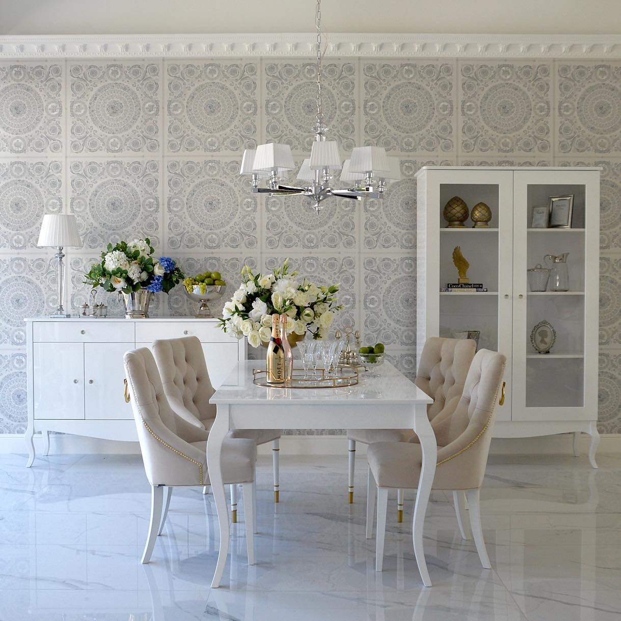 Elegancki stół Elena z białym blatem i krzesłami tapicerowanymi w beżowej tkaninie, z dekoracyjnymi złotymi nogami, na tle wzorzystej tapety i białych mebli.