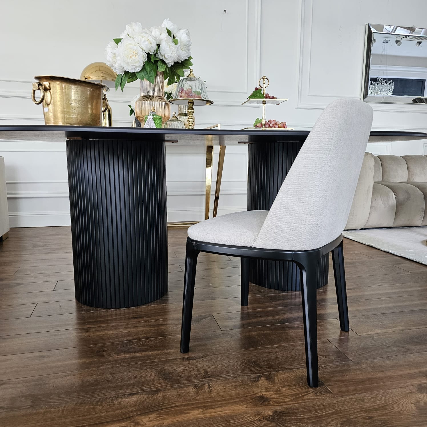 Eleganckie krzesło Napoli z jasną tapicerką i czarnymi nogami, ustawione przy stole z dekoracjami w nowoczesnym wnętrzu. Widoczny fragment sofy w tle.
