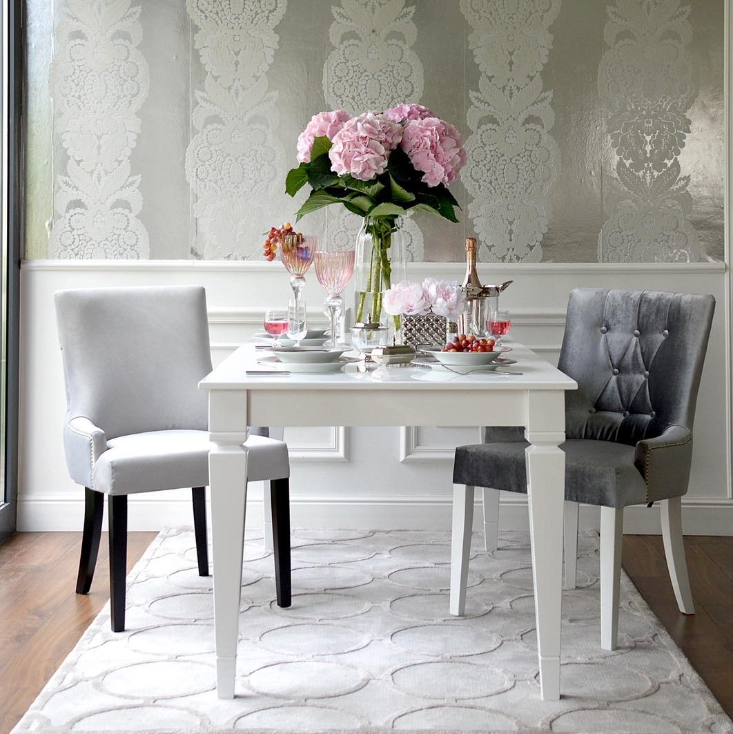 Elegancki stół z białym blatem i dwoma krzesłami (jasnoszarym i ciemnoszarym) na tle srebrnej tapety w kwiatowe wzory. Na stole zastawa, kwiaty i owoce.