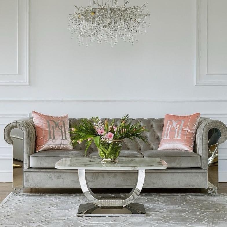 Szara sofa Chesterfield z lustrzanymi bokami, różowe poduszki z monogramem, marmurowy stolik kawowy i kryształowy żyrandol w eleganckim salonie.