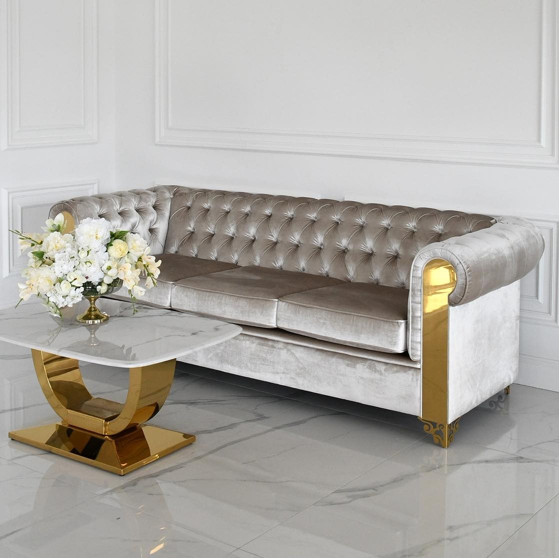 Ekskluzywna sofa Chesterfield w odcieniach szarości z pozłacanymi zdobieniami, ustawiona w minimalistycznym wnętrzu z marmurową podłogą i stolikiem kawowym.