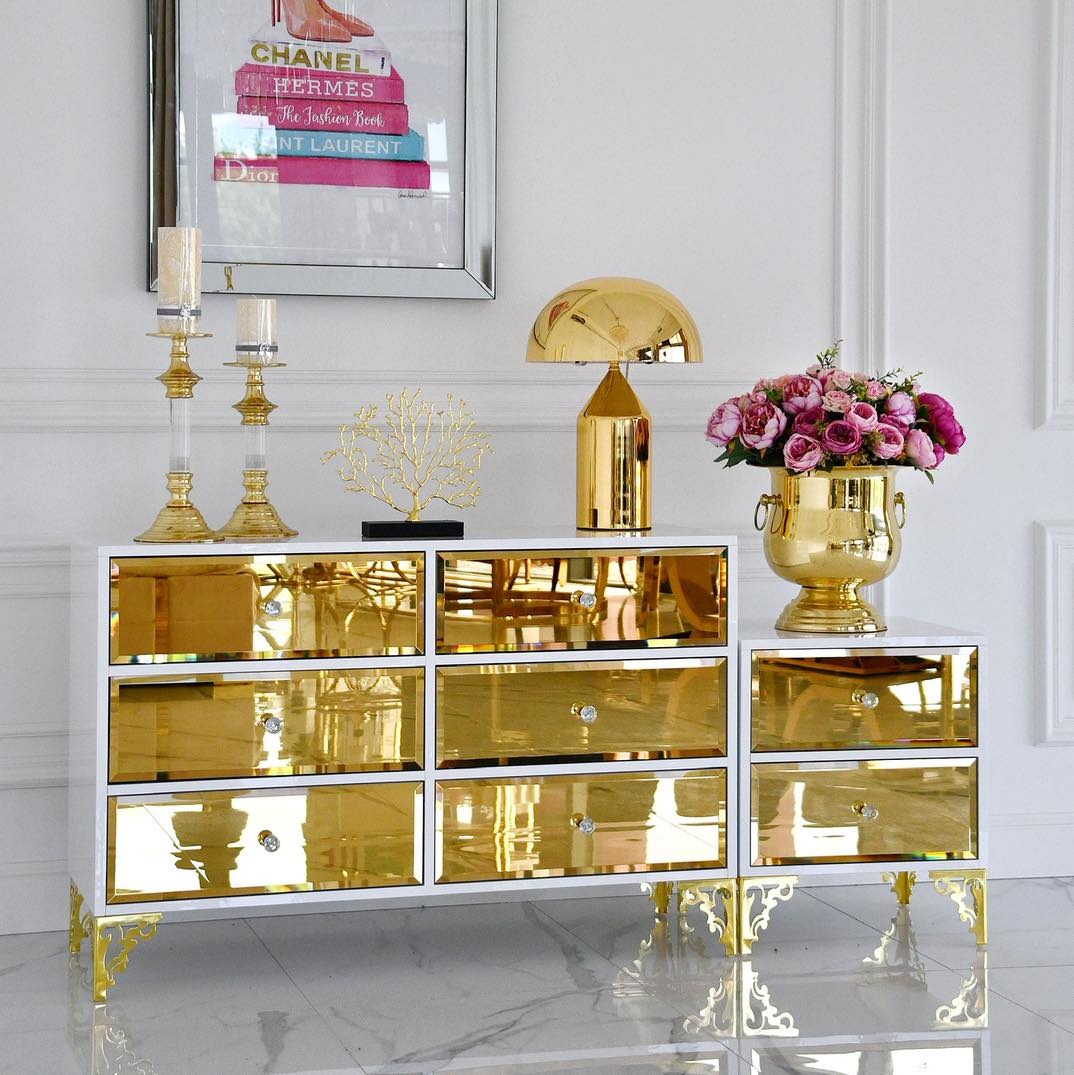 Biała komoda z dziewięcioma lustrzanymi szufladami i złotymi nóżkami, na blacie złota lampka, wazon z kwiatami i świeczniki. W tle obraz w srebrnej ramie.