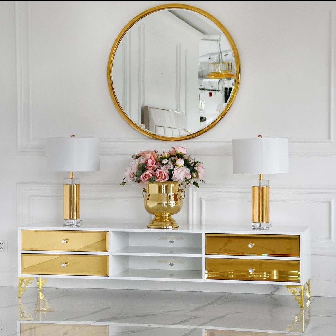 Biała komoda RTV z lustrzanymi, złotymi szufladami i zdobionymi nóżkami, złote lampy i lustro na tle białej ściany z dekoracyjnymi listwami.