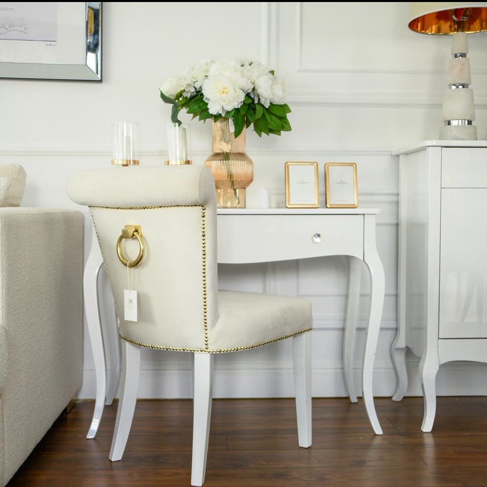 Tapicerowane krzesło z ozdobnym złotym uchwytem i nitami, ustawione przy białej toaletce z kwiatami i ramkami na zdjęcia w eleganckim, jasnym wnętrzu.