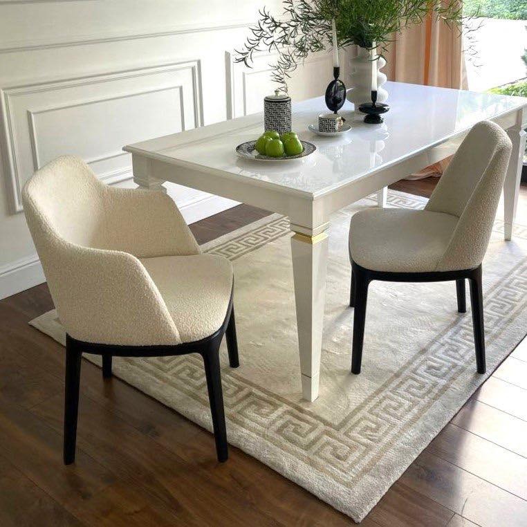 Biały stół z połyskiem i krzesła z beżową tapicerką boucle na wzorzystym dywanie. Dekoracje stołu: owoce, świeczniki. Eleganckie wnętrze z zasłonami w tle.