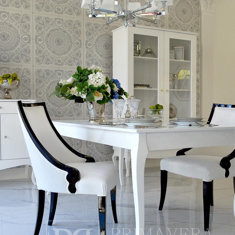 Eleganckie krzesło Carlotta z czarnymi zdobieniami i białym obiciem przy białym stole w jadalni w stylu glamour. W tle kredens i żyrandol.