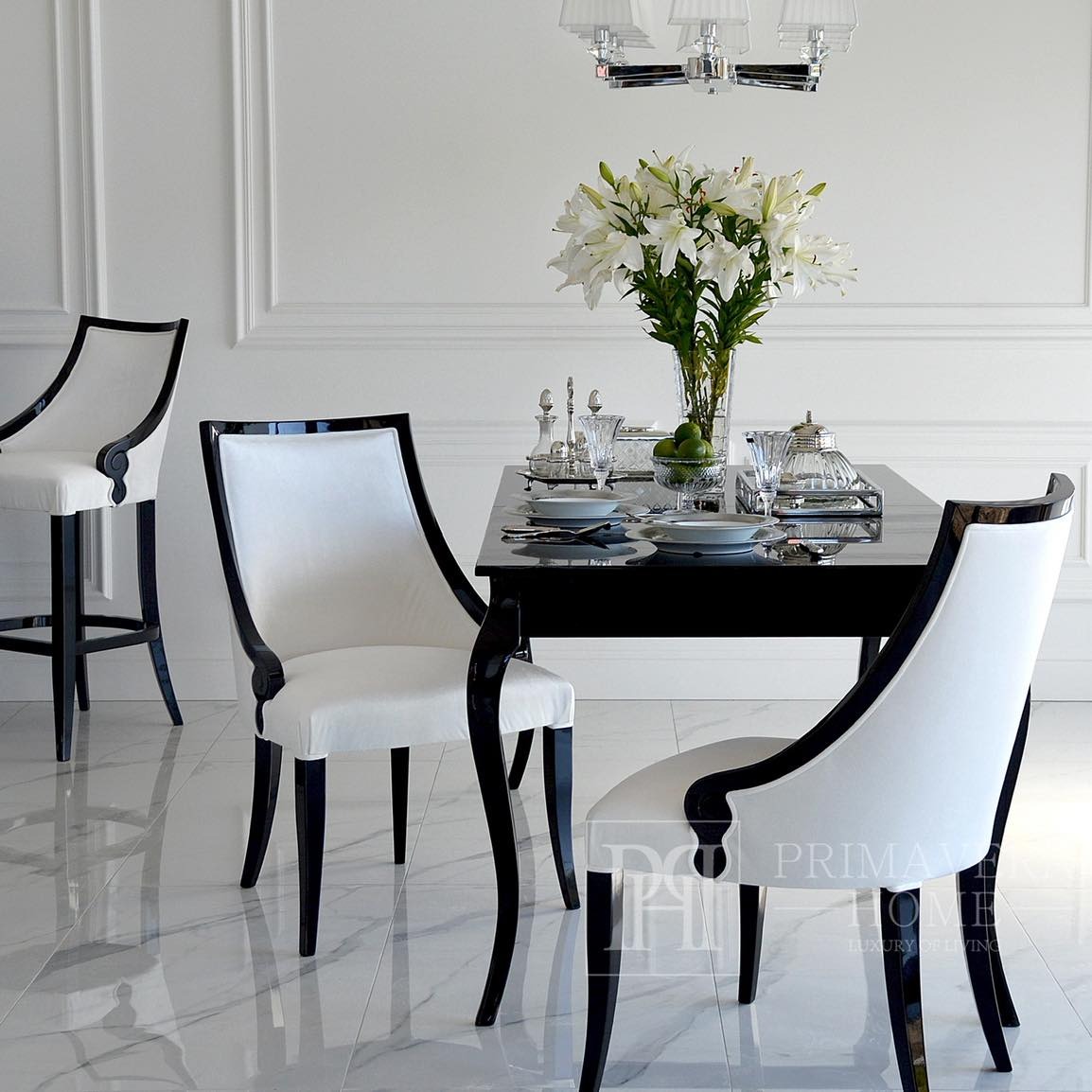 Elegancki stół z czarnym blatem i krzesłami tapicerowanymi na biało z czarnymi, giętymi nogami, uzupełniony bukietem lilii i srebrną zastawą w jasnym wnętrzu.