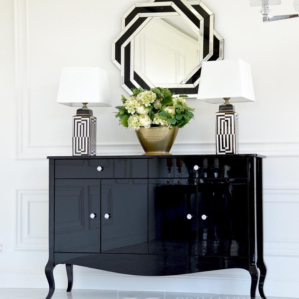 Czarna, lakierowana komoda w stylu glamour z giętymi nogami i kryształowymi uchwytami, na niej dekoracje: dwie lampy i bukiet kwiatów. Nad komodą lustro.