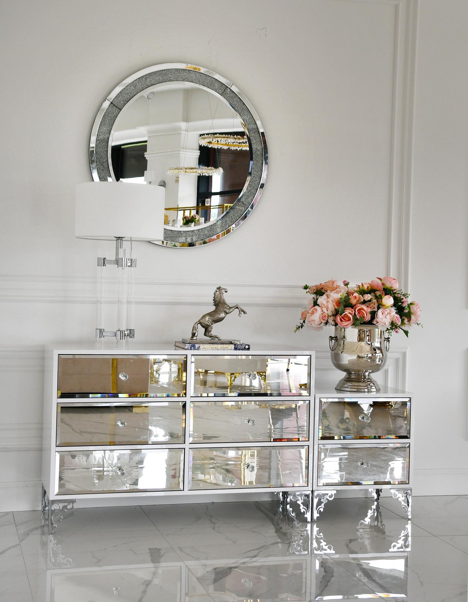 Komoda Venice z lustrzanymi frontami i srebrnymi zdobieniami, na blacie lampa, figurka konia i wazon z różowymi różami. Nad komodą okrągłe lustro.