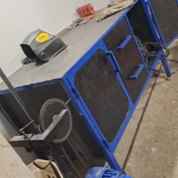 Stół roboczy z niebieską ramą, maską spawalniczą Stanley, szlifierką, akcesoriami, rękawicami i silnikiem elektrycznym w warsztacie, lekko obrócony w lewo.