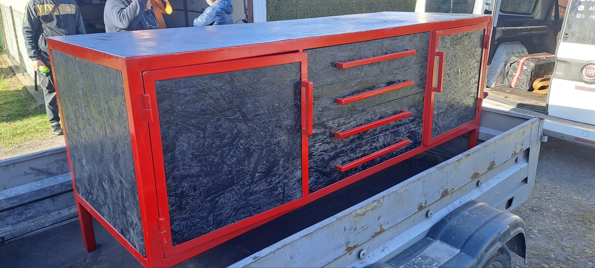 Metalowa szafka z trzema szufladami i drzwiczkami, obudowana czerwoną ramą, umieszczona na przyczepie. Czarny wzór płyty meblowej.