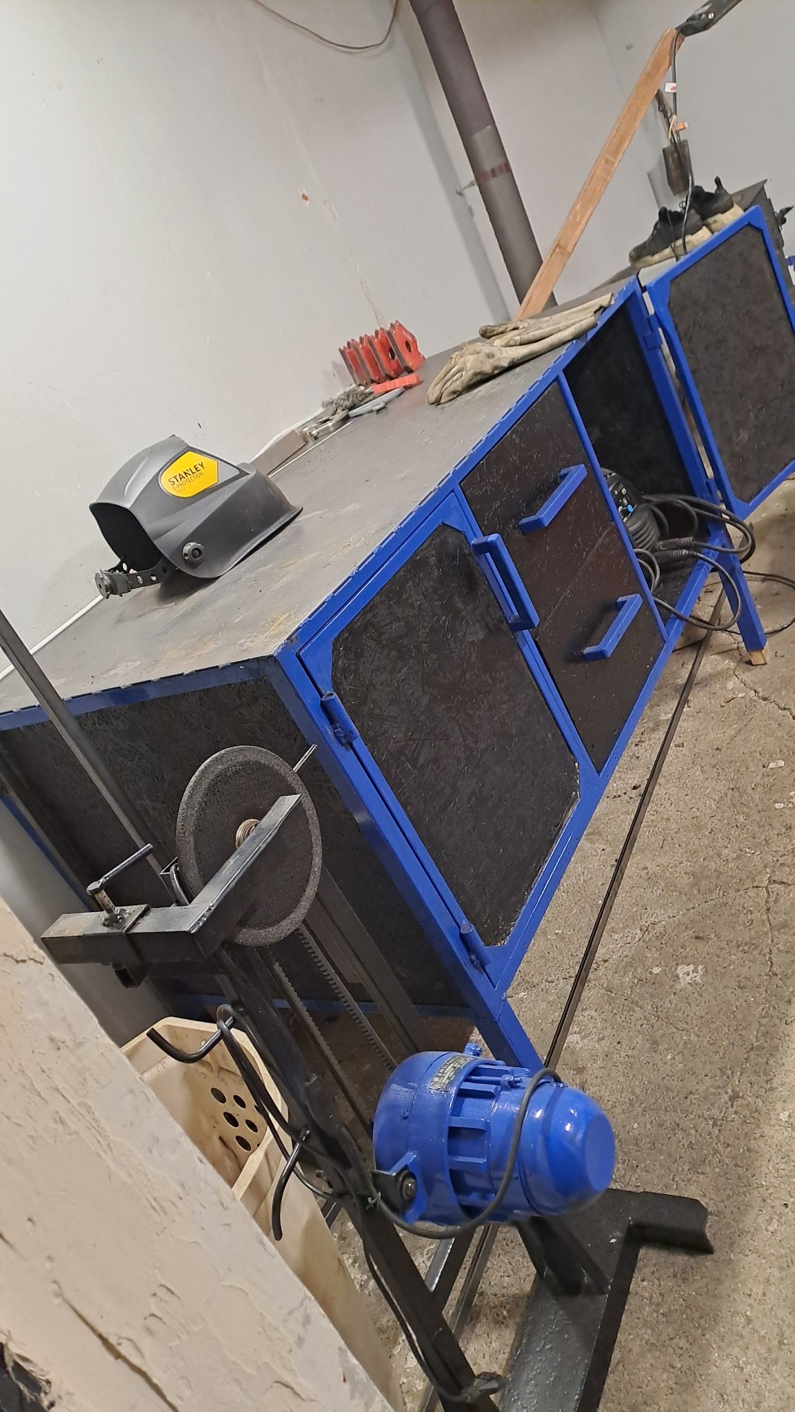 Stół roboczy z niebieską ramą, maską spawalniczą Stanley, szlifierką, akcesoriami, rękawicami i silnikiem elektrycznym w warsztacie, lekko obrócony w lewo.