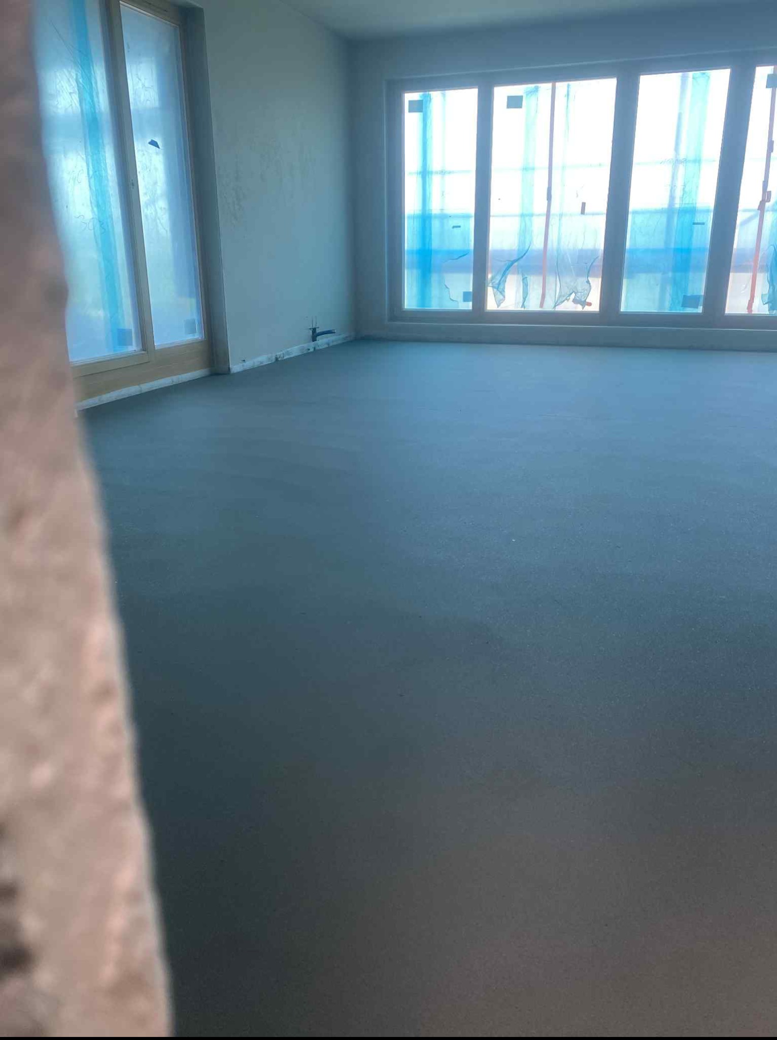 Gładka, szara posadzka jastrychowa w jasnym pomieszczeniu z oknami zabezpieczonymi folią. Świeżo wylana podłoga w stanie surowym.