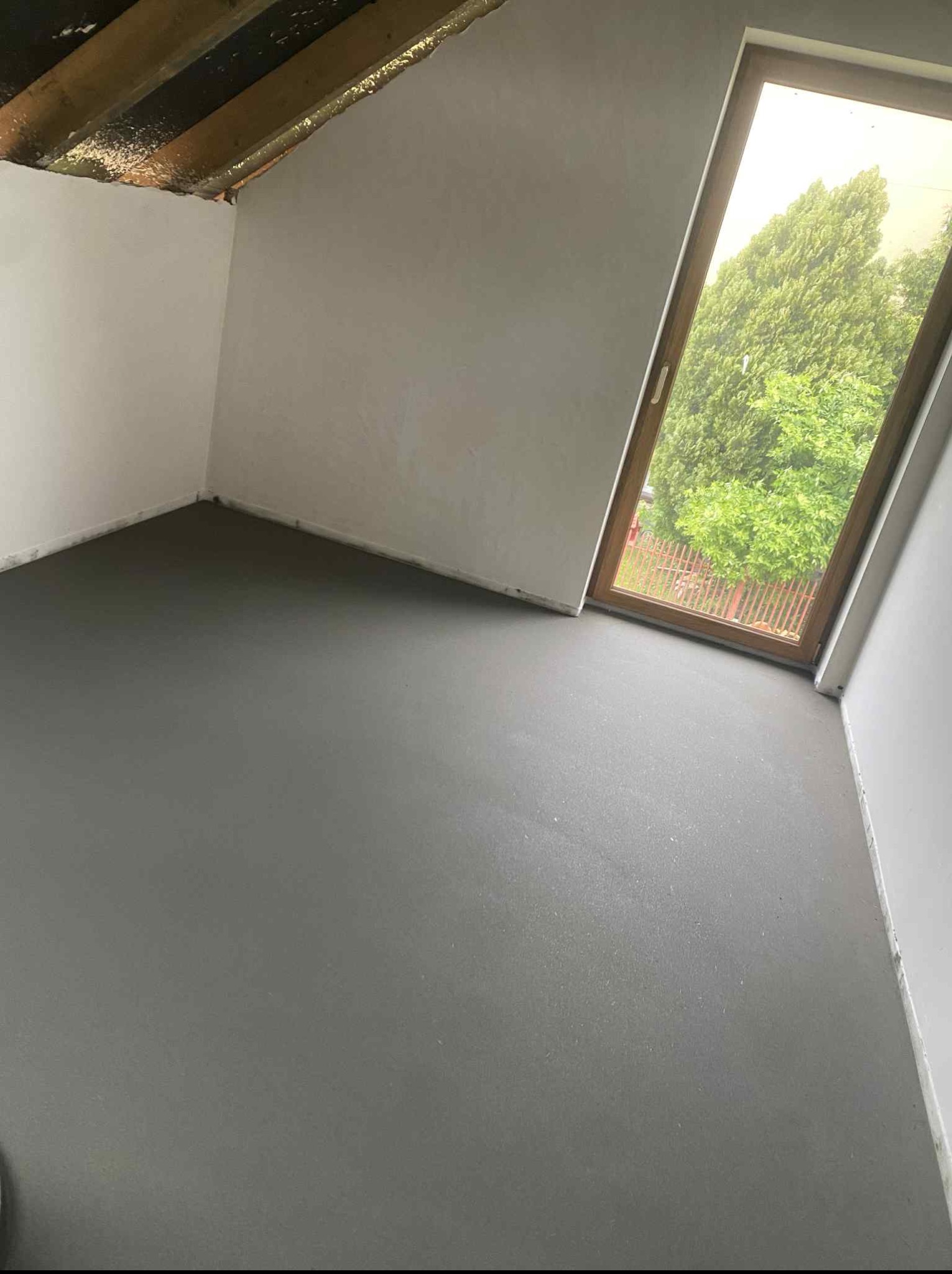 Świeżo wylana, gładka posadzka jastrychowa w jasnym pokoju na poddaszu z oknem dachowym, przez które widać zieleń drzew. Szara, jednolita powierzchnia podłogi.