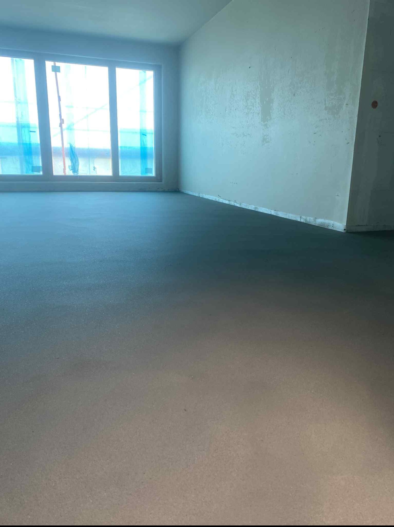 Świeżo wylana, gładka posadzka jastrychowa w jasnym pomieszczeniu z dużym oknem, na którym widoczne są rusztowania. Wnętrze w stanie surowym.