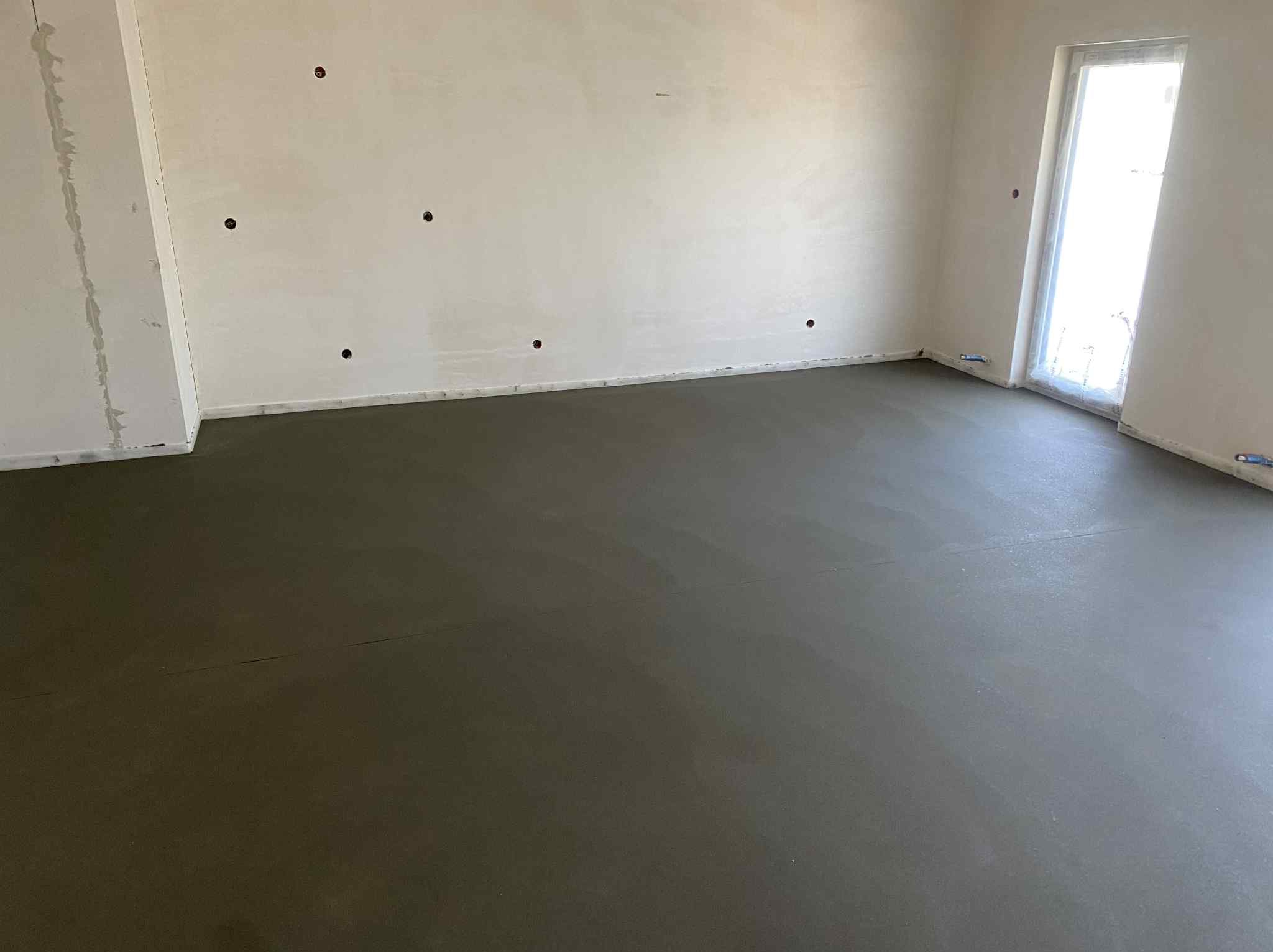 Świeżo wylana, gładka posadzka jastrychowa w jasnym pomieszczeniu z oknem. Widoczne otwory w ścianie i ślady po łatach gipsowych.