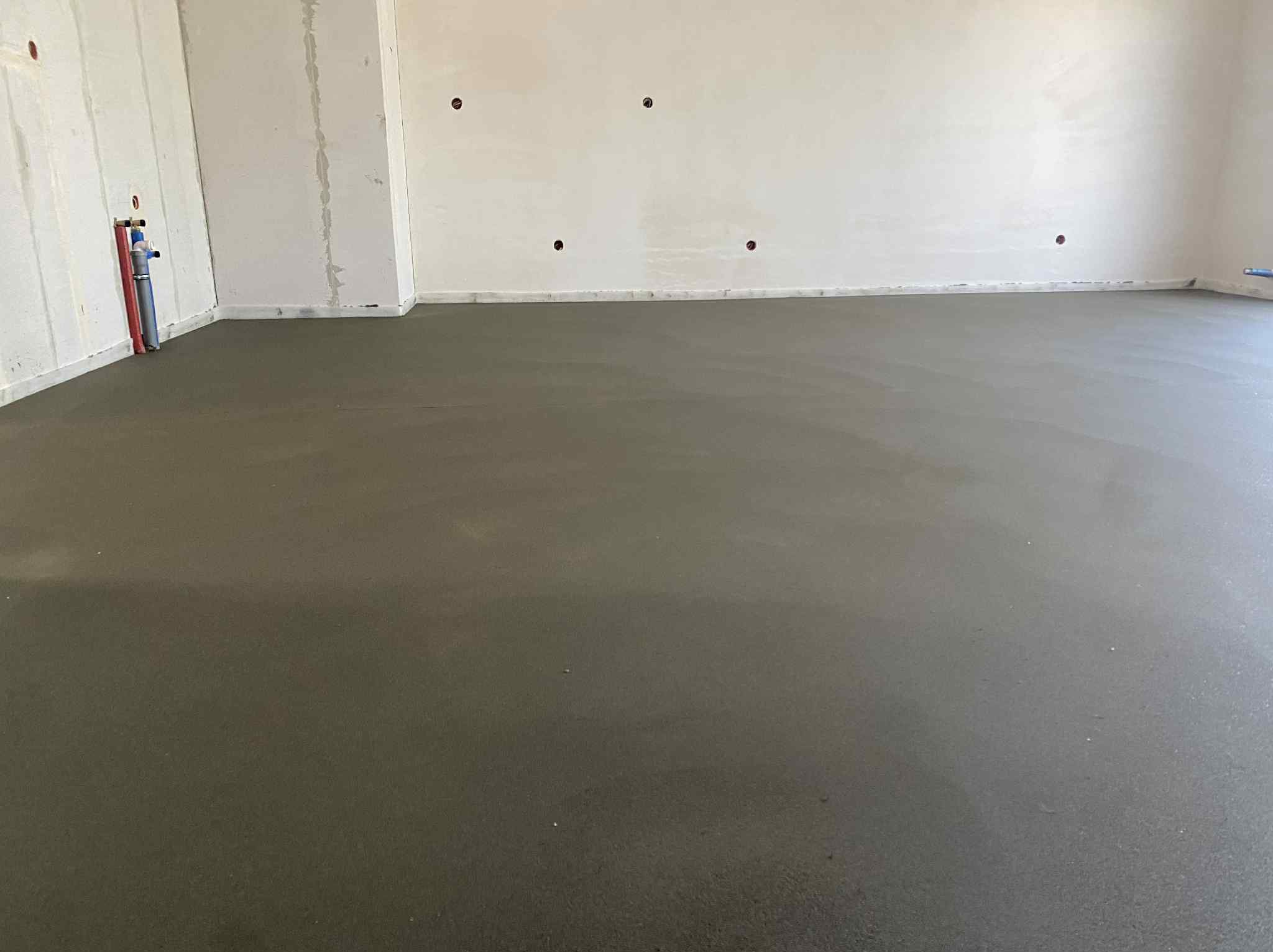 Gładka, szara posadzka jastrychowa w surowym wnętrzu. Widoczne rury instalacyjne przy ścianie i otwory w ścianie. Minimalistyczny, nowoczesny styl.