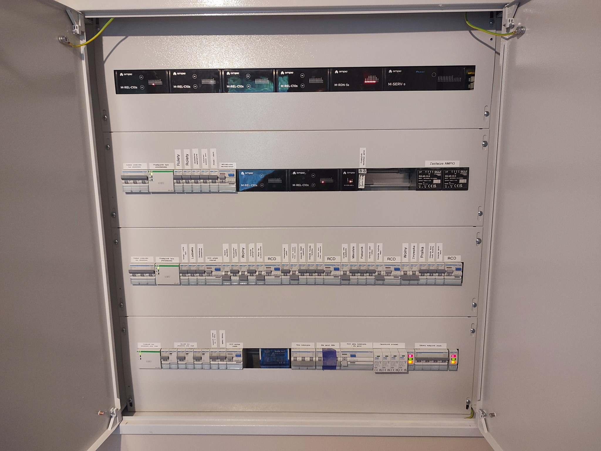 Rozdzielnica elektryczna z modułami firmy Ampio, widoczne bezpieczniki, wyłączniki RCD i moduły sterujące. Otwarta obudowa, kable ochronne.