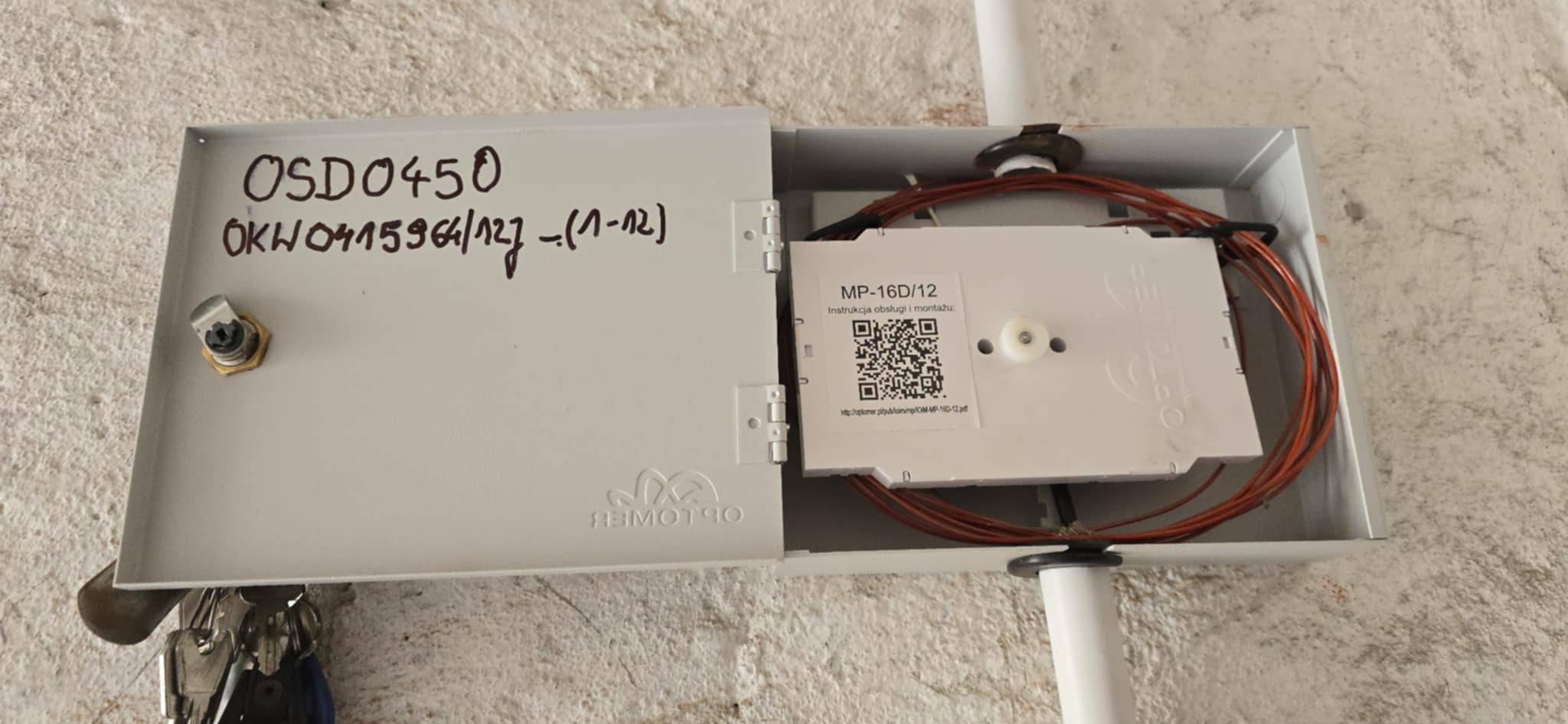 Otwarta skrzynka telekomunikacyjna z kablem światłowodowym wewnątrz, przymocowana do ściany. Widoczne oznaczenia OSD0450 i MP-16D/12.