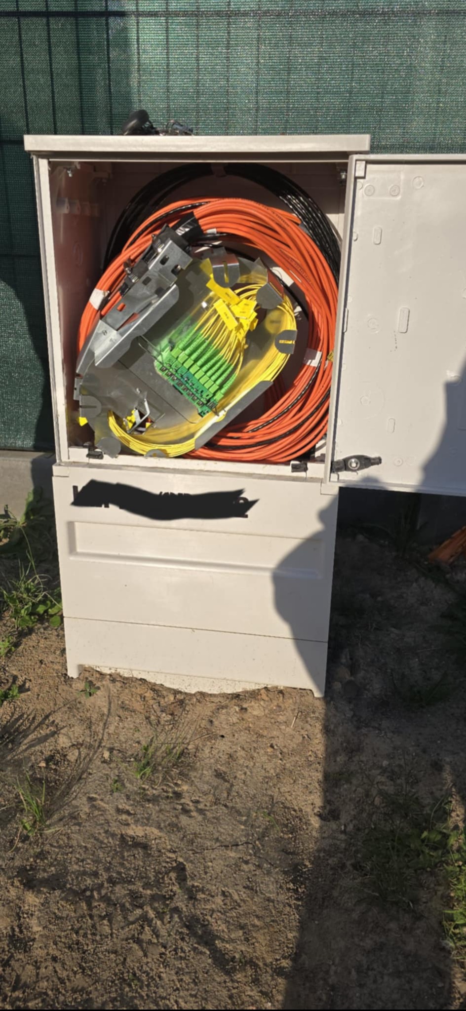 Otwarta szafka telekomunikacyjna z pomarańczowymi i żółto-zielonymi kablami światłowodowymi, umieszczona na ziemi, z zielonym płotem w tle.