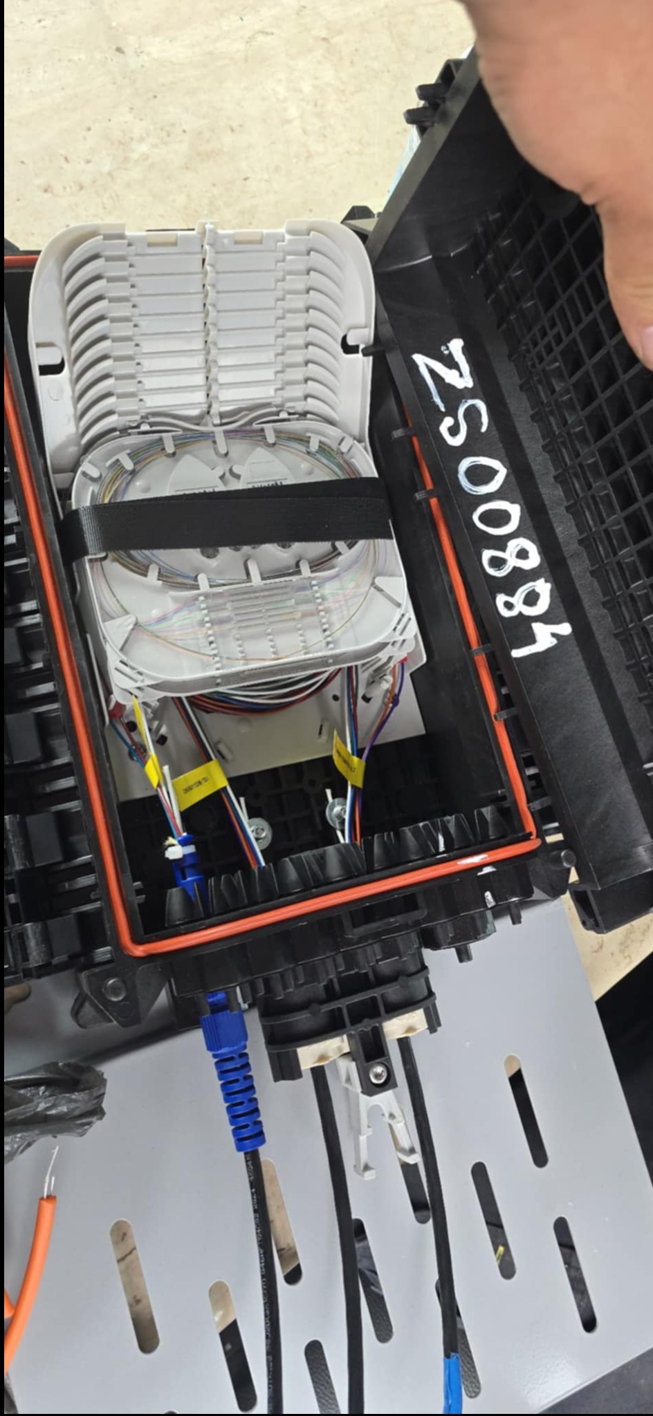 Otwarta skrzynka telekomunikacyjna z ułożonymi kablami światłowodowymi, zabezpieczonymi czarnym paskiem. Widoczne kable z niebieskimi końcówkami i oznaczenia na obudowie.