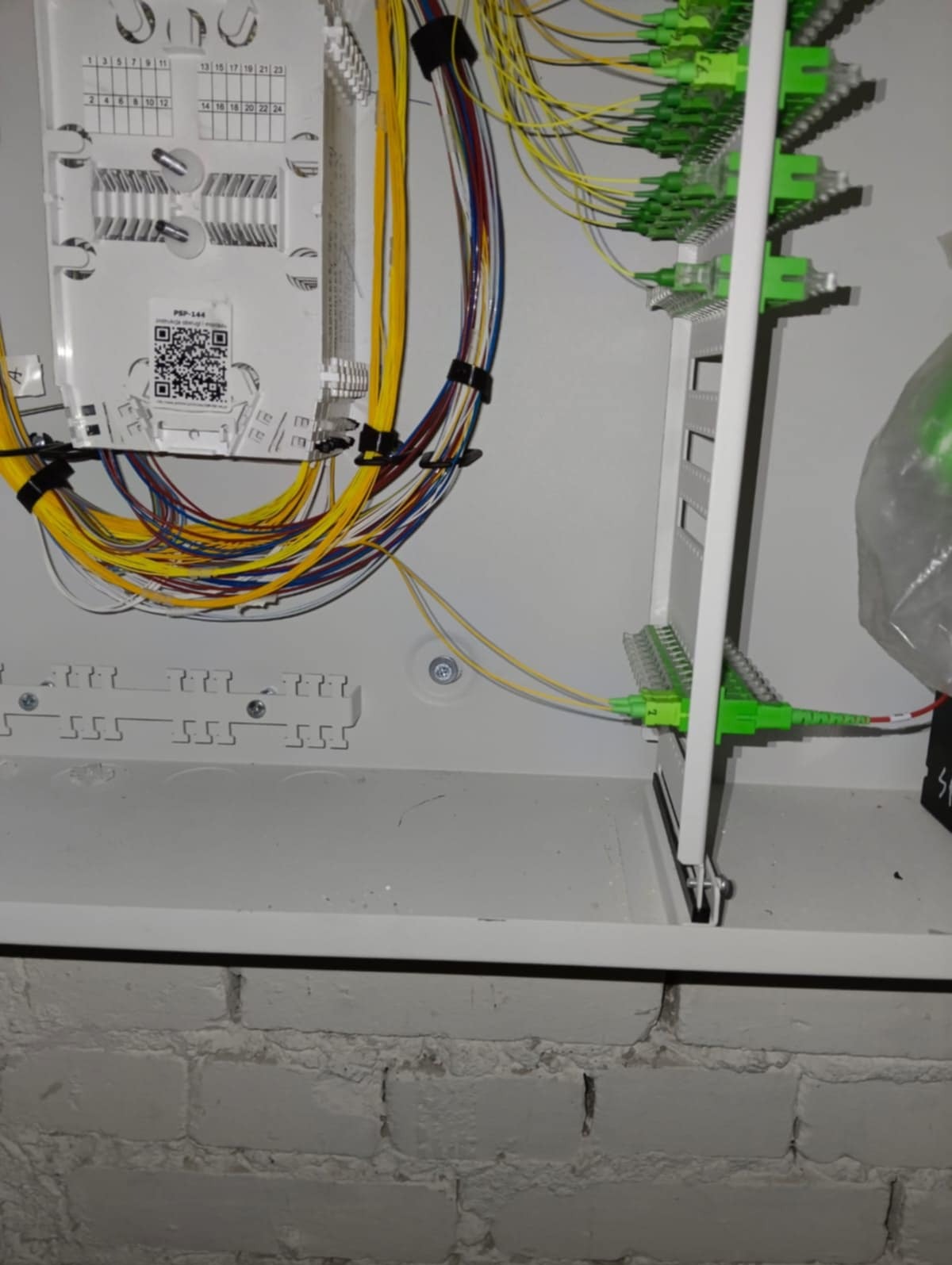 Rozdzielnica światłowodowa z podłączonymi kablami w kolorach żółtym, niebieskim i zielonym. Widoczna biała obudowa i fragment ściany z cegły.