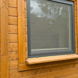 Moskitiera ramkowa w oknie drewnianego domu z widocznym refleksem drzew w siatce. Szara rama moskitiery kontrastuje z ciepłym odcieniem drewna elewacji.