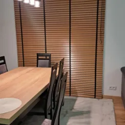 Drewniane żaluzje poziome w odcieniach brązu, zamontowane w oknie jadalni w Końskich. Widoczne czarne linki sterujące i fragment stołu z krzesłami.