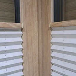 Zamknięte, białe plisy okienne na drewnianej ramie okiennej. Widoczne detale plisowania i sznurki. Ujęcie z bliska, podkreślające strukturę materiału i precyzję montażu.