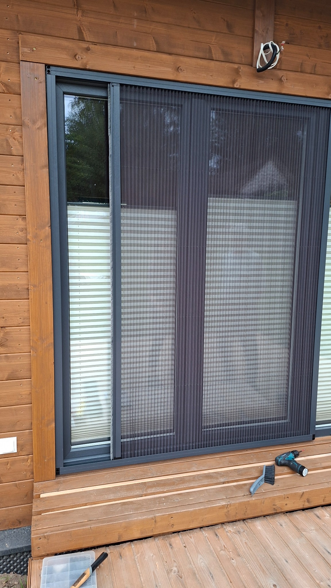 Zamontowana moskitiera plisowana w oknie drewnianego domu. Widoczne narzędzia montażowe, plisy wewnątrz okna. Całość w naturalnej kolorystyce drewna i szarości.